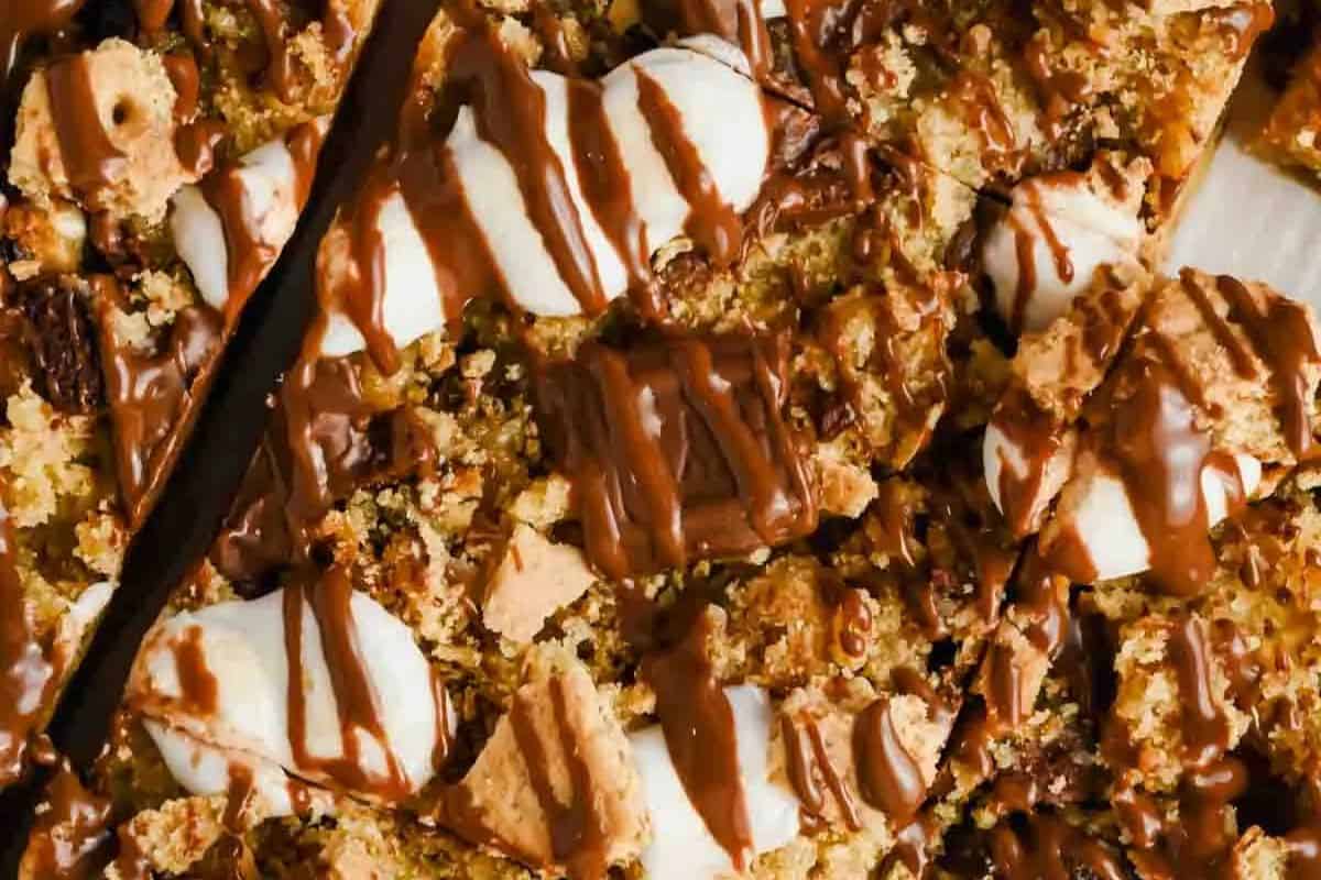  A top-down view of a s'mores casserole or bar dessert, cut into pieces. It's covered in crumbled graham crackers, melted marshmallows, and drizzled with melted chocolate.