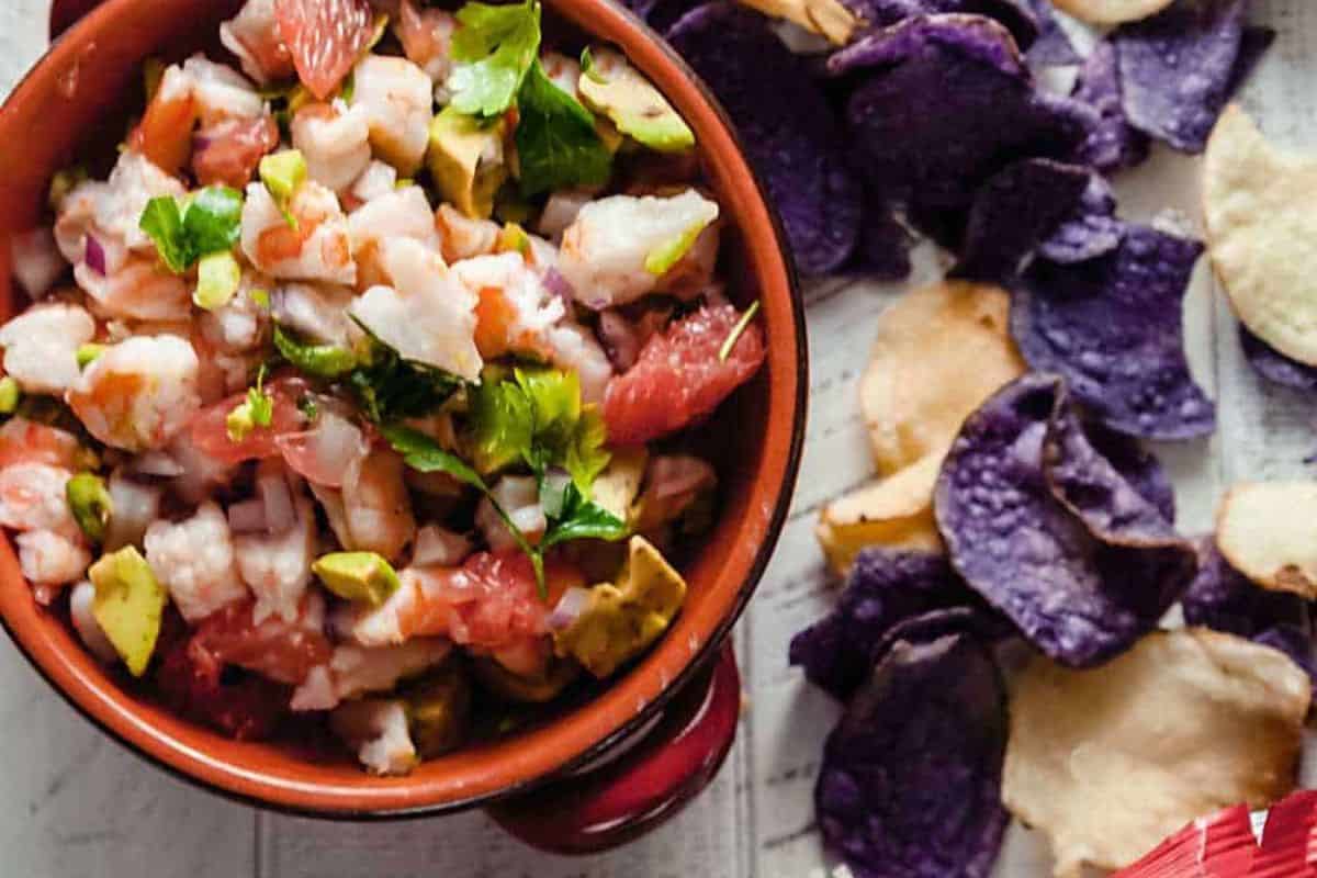 A top-down shot of a bowl of ceviche with shrimp, avocado, red onion, and pink grapefruit segments, garnished with fresh herbs. Next to the bowl are scattered purple and light-colored potato chips.