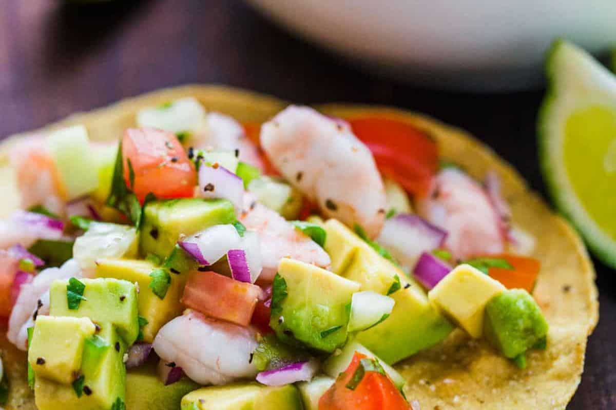 A close-up of a tostada topped with shrimp, diced avocado, tomato, red onion, cucumber, and cilantro, served with fresh lime in the background—perfect for those seeking vibrant new recipes.