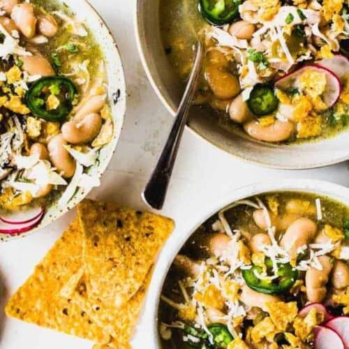 Three bowls of white bean chili garnished with jalapeño slices, radishes, shredded cheese, and crushed tortilla chips, served with extra chips and a wedge of fresh lime—perfect for trying new recipes or cooking up a cozy meal.