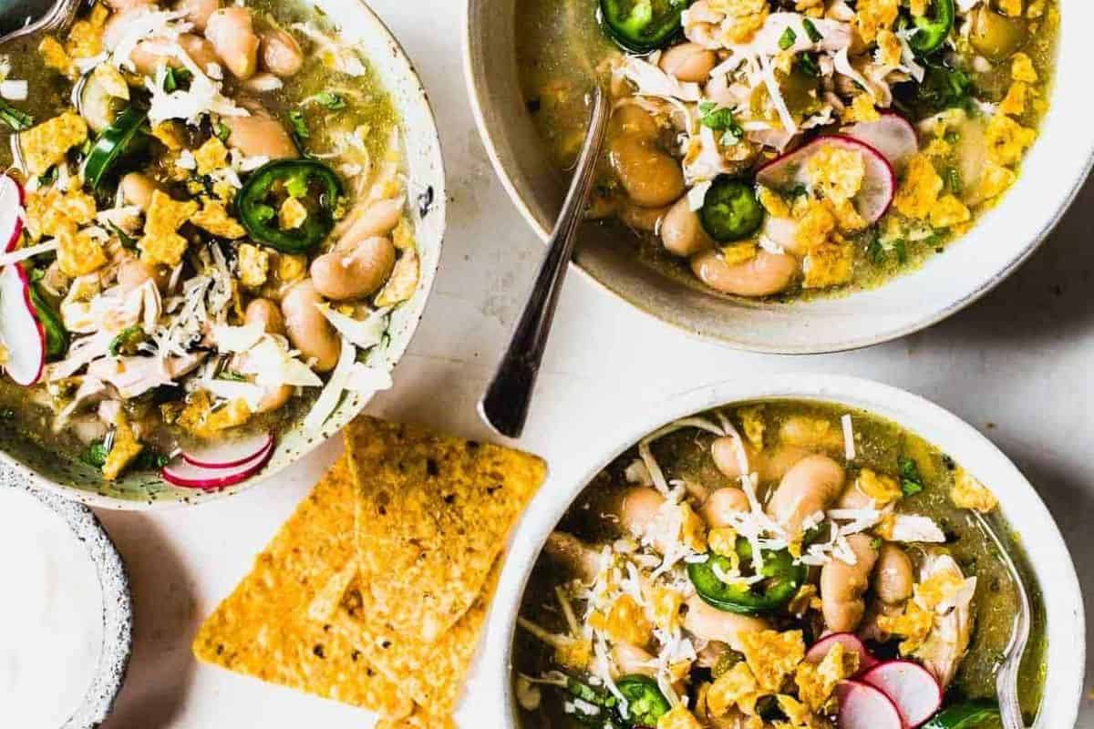 Three bowls of white bean chili garnished with jalapeño slices, radishes, shredded cheese, and crushed tortilla chips, served with extra chips and a wedge of fresh lime—perfect for trying new recipes or cooking up a cozy meal.