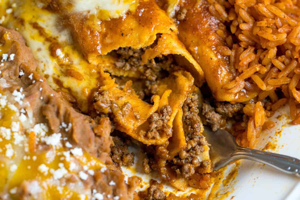 Close-up of a plate with ground beef enchiladas topped with sauce, refried beans made with canned beans and sprinkled with cheese, plus a serving of Mexican rice—perfect inspiration for delicious dishes and new recipes.