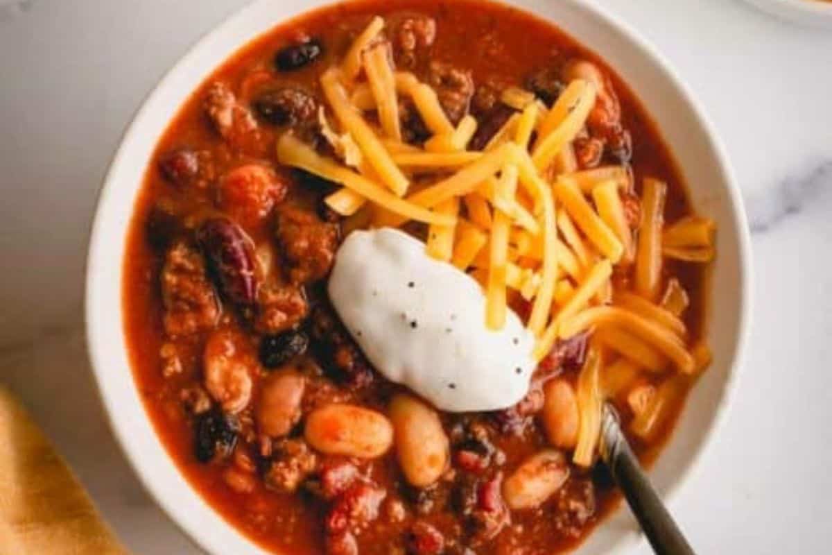 A comforting bowl of chili with ground meat, beans, shredded cheddar cheese, and a dollop of sour cream makes the perfect dinner. A spoon rests inside, ready to enjoy this classic take on beef soup recipes.