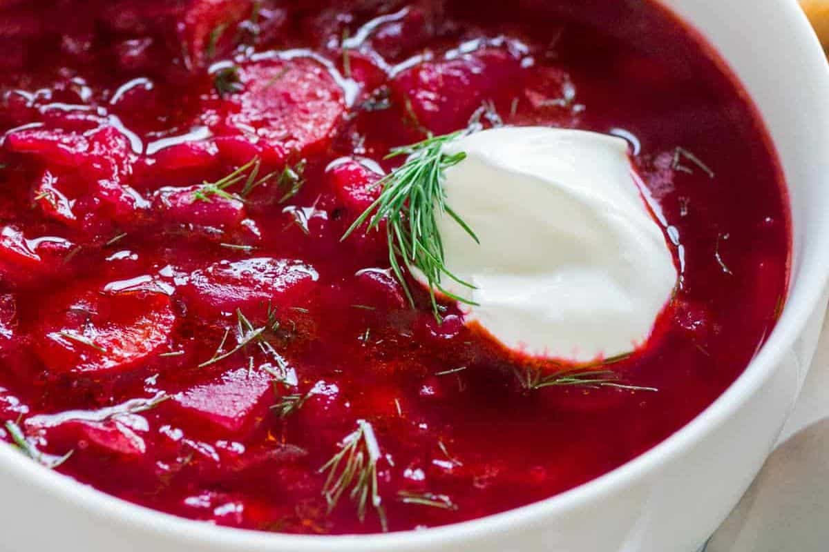 A close-up of a bowl of classic red borscht (beet soup) with pieces of beet and other vegetables, garnished with a generous dollop of white sour cream and a sprig of fresh dill.