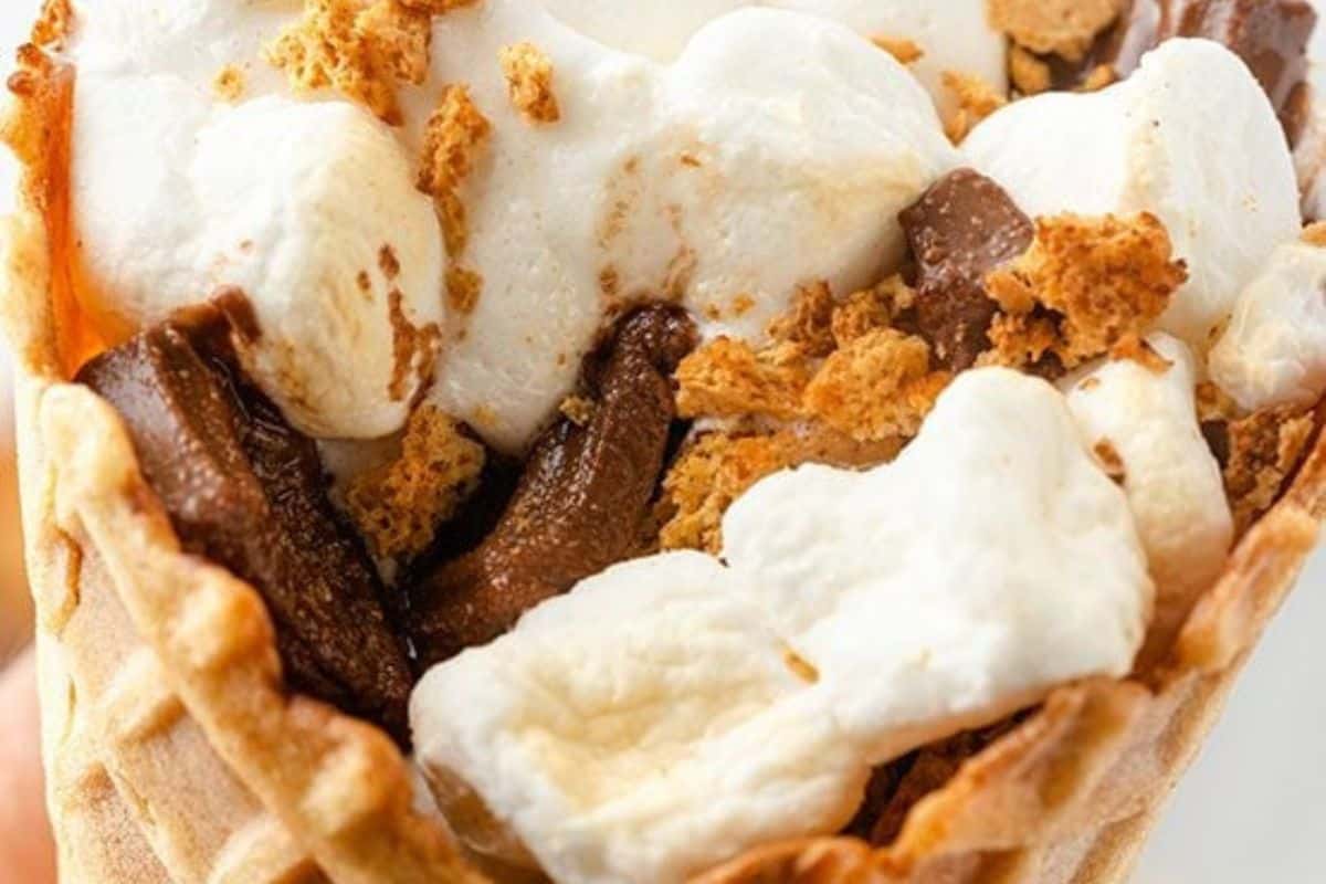 A close-up shot inside a waffle cone filled with s'mores ingredients. There are large pieces of white marshmallow, chunks of chocolate, and golden-brown graham cracker crumbs visible within the cone.