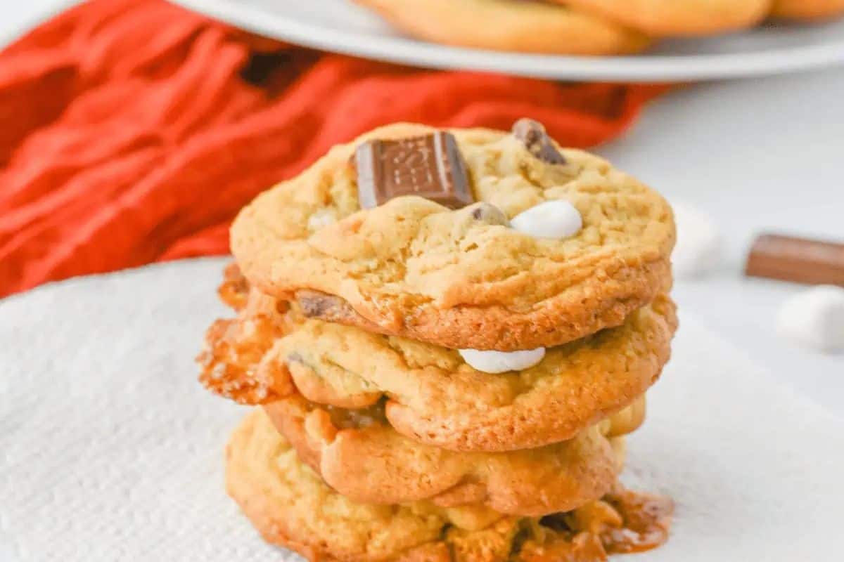 A stack of three golden-brown s'mores cookies on a white napkin, with a red napkin in the background. Each cookie has visible chocolate chips, pieces of chocolate bar, and white mini marshmallows.