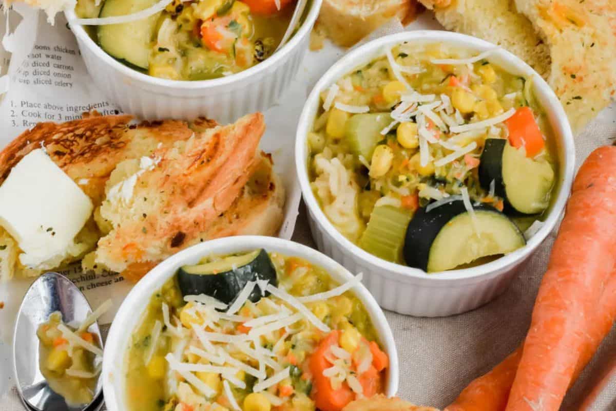 Three small bowls of vegetable soup, a classic in Dutch oven recipes, feature zucchini, carrots, corn, and shredded cheese, surrounded by slices of bread, butter, and fresh carrots on a light surface.