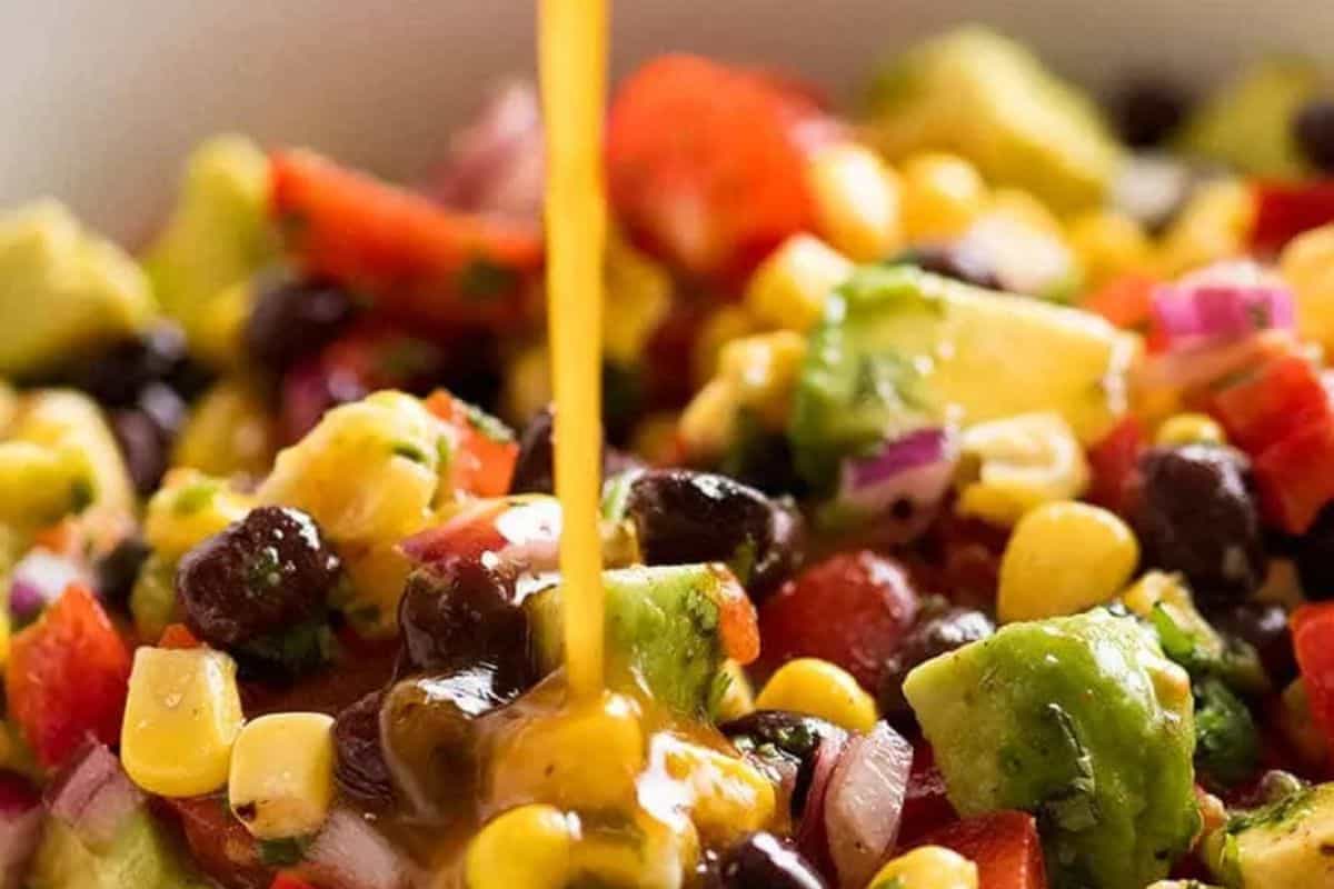  A close-up of cowboy caviar or a similar bean salad with corn, black beans, red onion, and avocado, with a stream of dressing being poured over it.