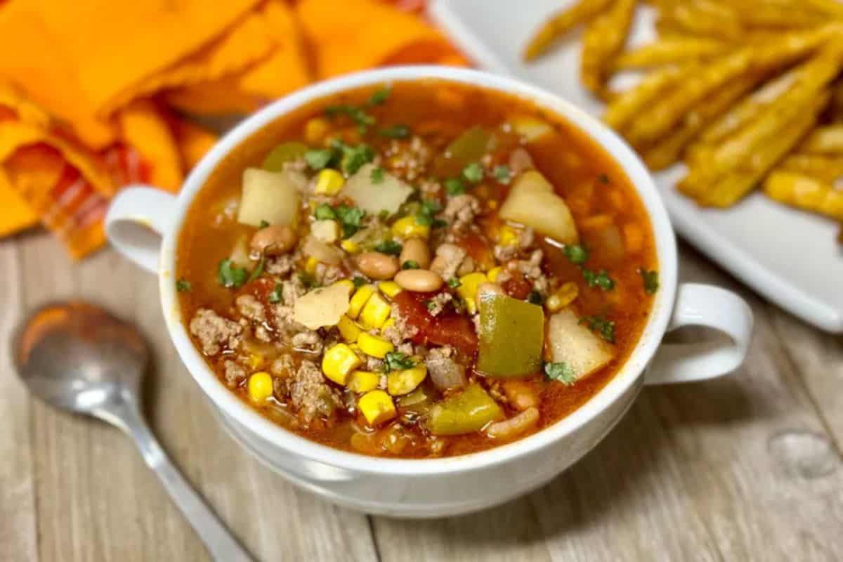 A white bowl of comforting vegetable and ground meat soup—with potatoes, corn, beans, and herbs—sits on a wooden table next to a spoon and fries. Perfect for your dinner rotation or anyone looking for hearty beef soup recipes.