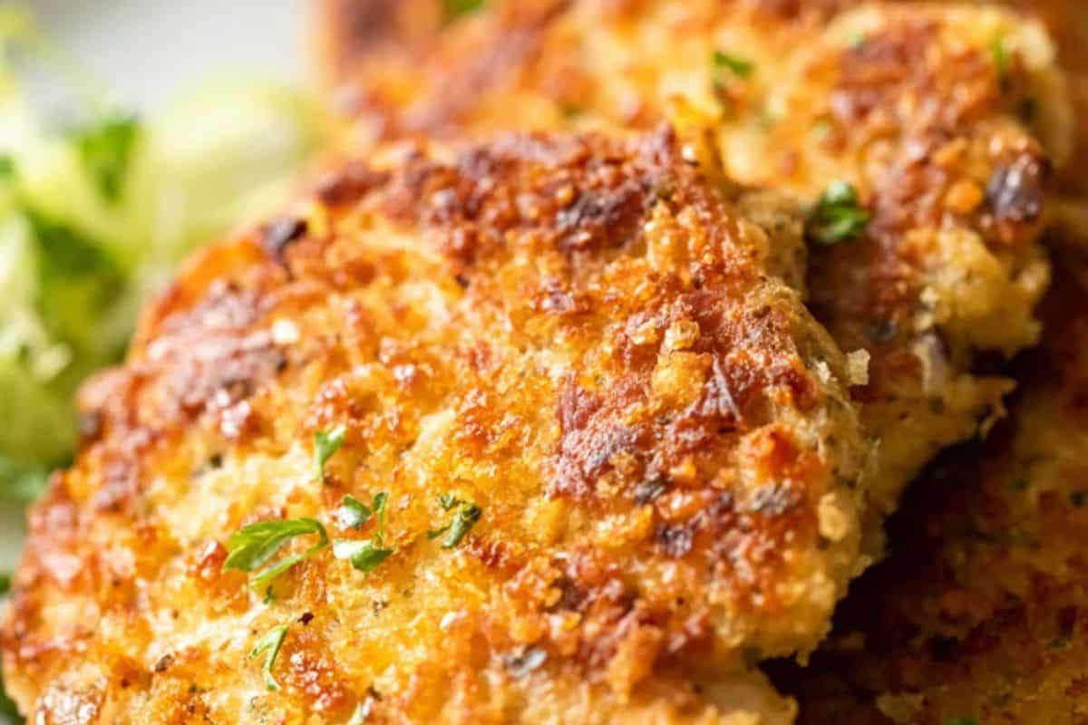 Close-up of golden-brown, crispy salmon patties garnished with chopped herbs—an inspiration for easy tuna meals or creative tuna recipes, with a blurred side of vegetables in the background.