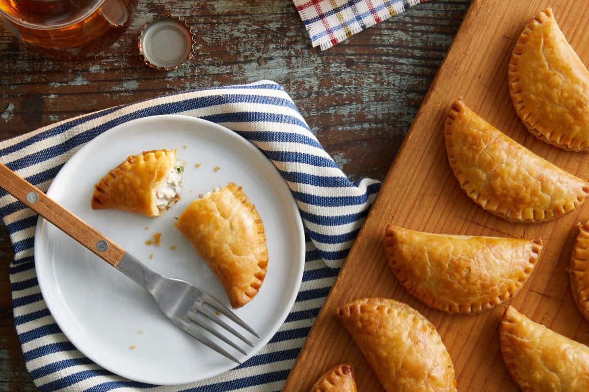 A plate with a half-eaten savory pastry and a fork sits on a striped cloth; several whole pastries, perfect for easy tuna meals, are arranged on a wooden board beside it.
