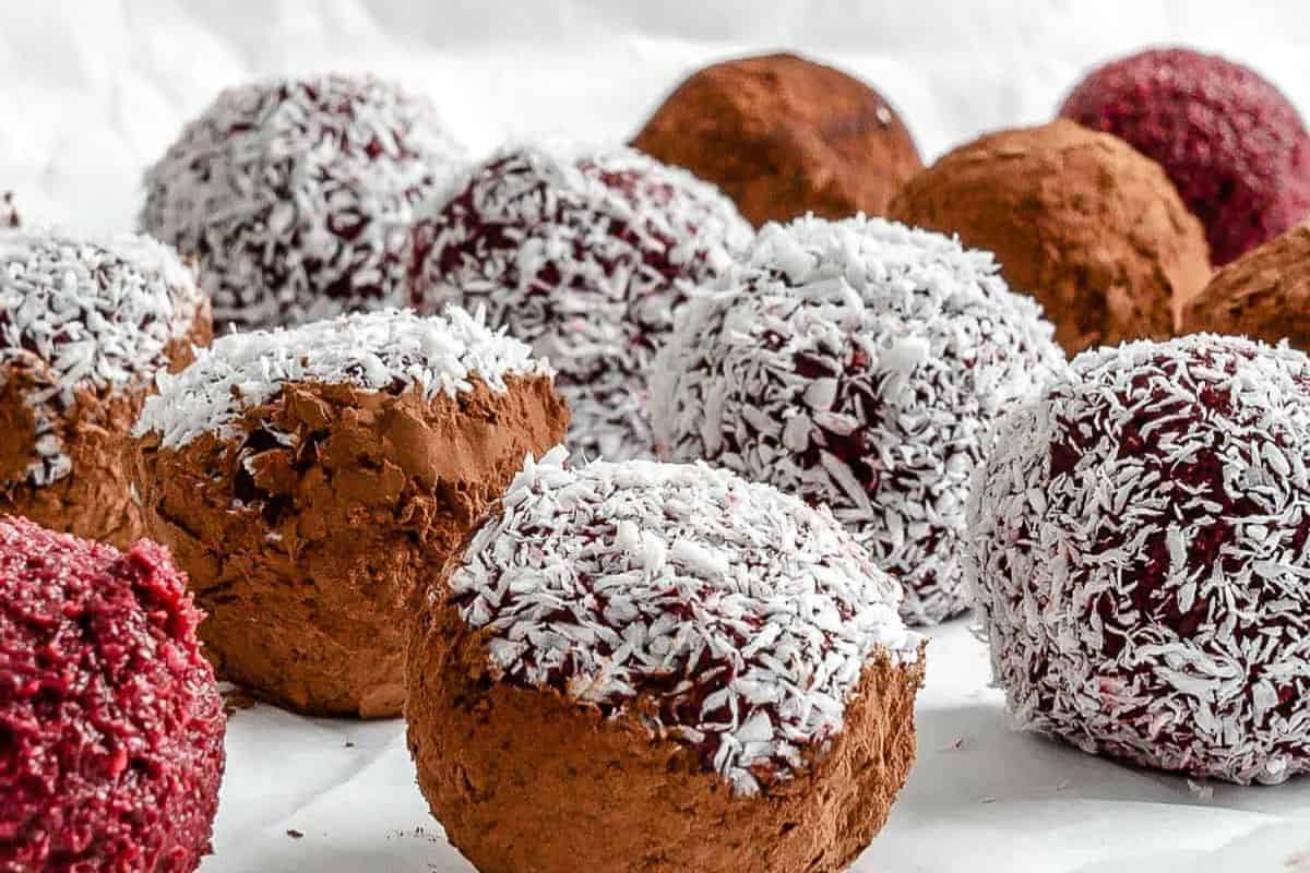  A close-up of numerous round energy balls or truffles. Some are coated in shredded white coconut, others in brown cocoa powder, and some in a reddish powder (possibly beet powder), arranged on a white surface.