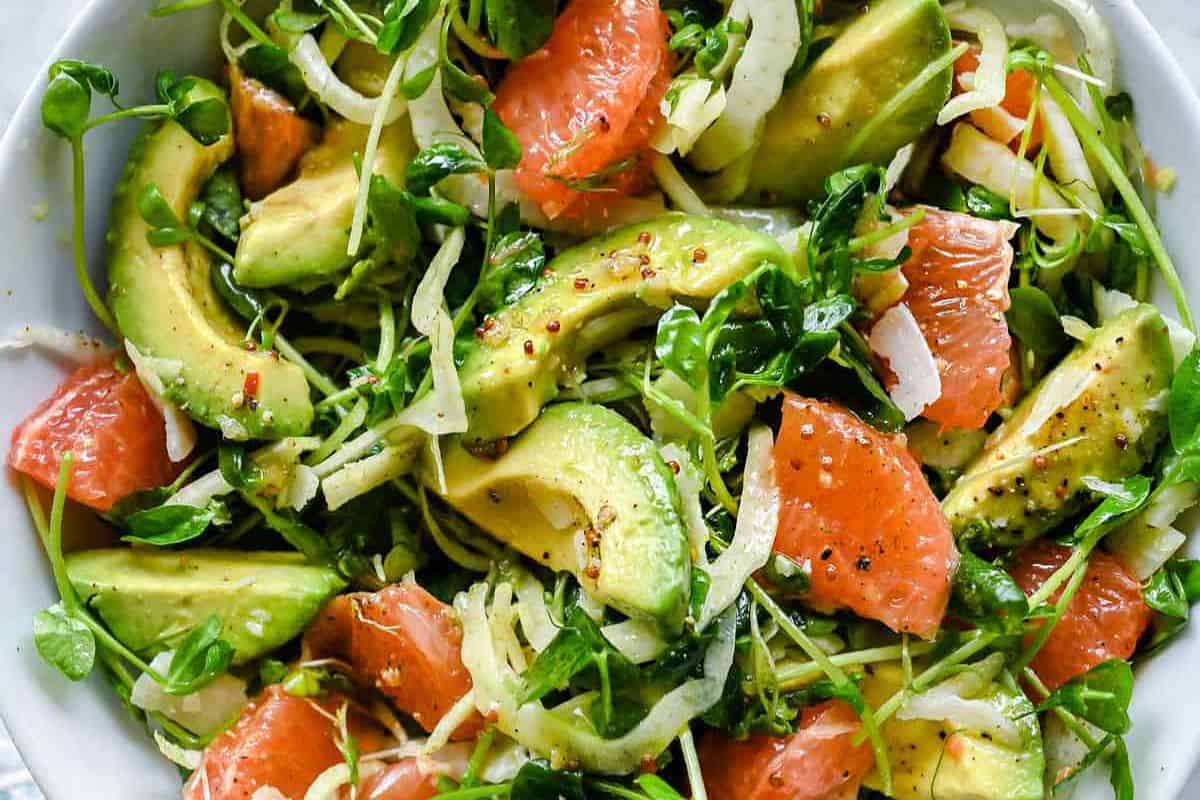 A salad with sliced avocado, grapefruit segments, shredded greens, and sprouts, lightly seasoned with black pepper and dressing—perfect for those seeking refreshing grapefruit recipes.
