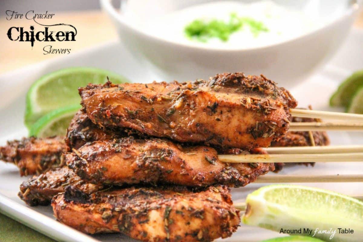 Grilled chicken skewers with herbs and spices are stacked on a plate, served with lime wedges and a bowl of white dipping sauce—perfect for party food recipes that everyone will love.