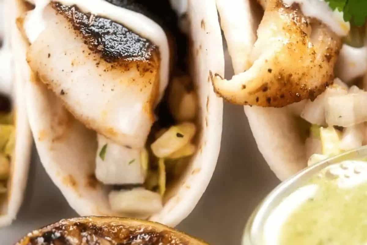 Close-up of fish tacos with grilled fish, diced onions, and sauce in soft tortillas, served with a slice of fresh lime and a side of green sauce—perfect for anyone who loves delicious cooking or experimenting with new recipes.