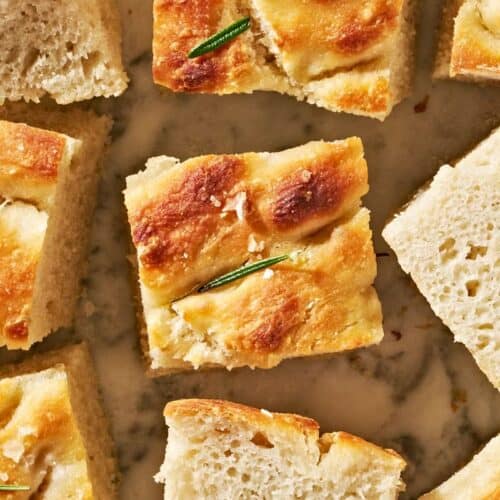 Close-up of several square pieces of golden-brown focaccia bread, some topped with rosemary, on a light surface—a must try for fans of quick breads you can bake anytime.