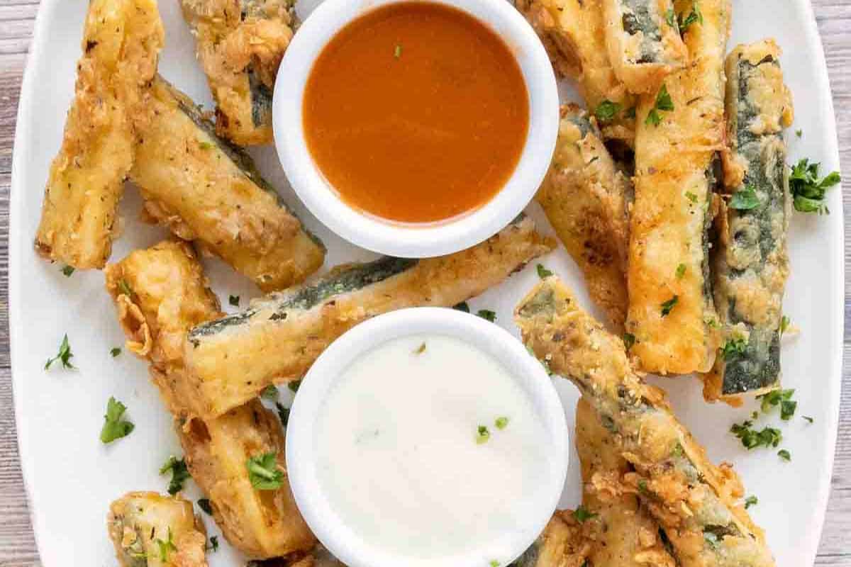 Plate of battered and fried zucchini sticks garnished with parsley, served with two dipping sauces—one orange, one white—making for a crunchy twist on classic cold appetizer ideas.