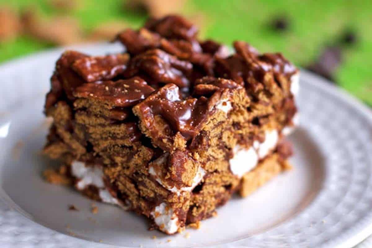 A square s'mores bar on a white plate, featuring a base layer topped with white marshmallows, chocolate, and a generous amount of Golden Grahams cereal, all drizzled with chocolate.