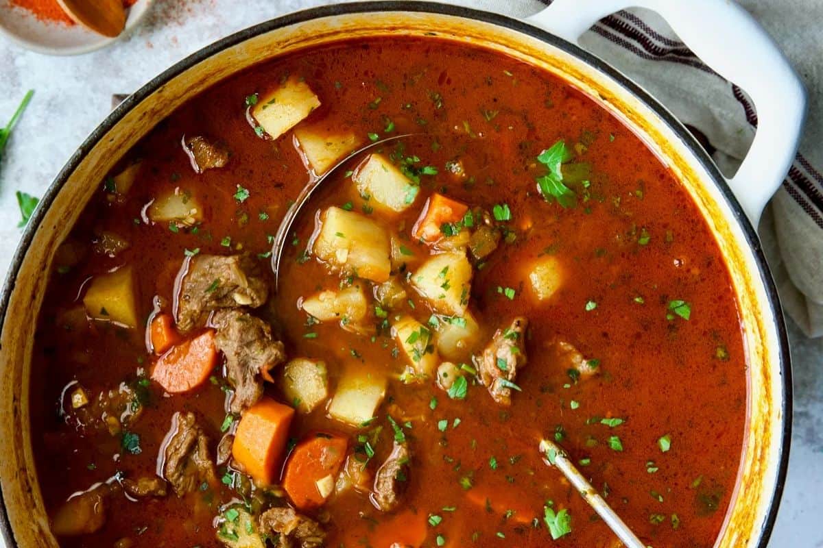 A pot of comforting soup with chunks of beef, potatoes, carrots, and fresh herbs in a rich, red broth, with a spoon resting inside—perfect for your dinner rotation.
