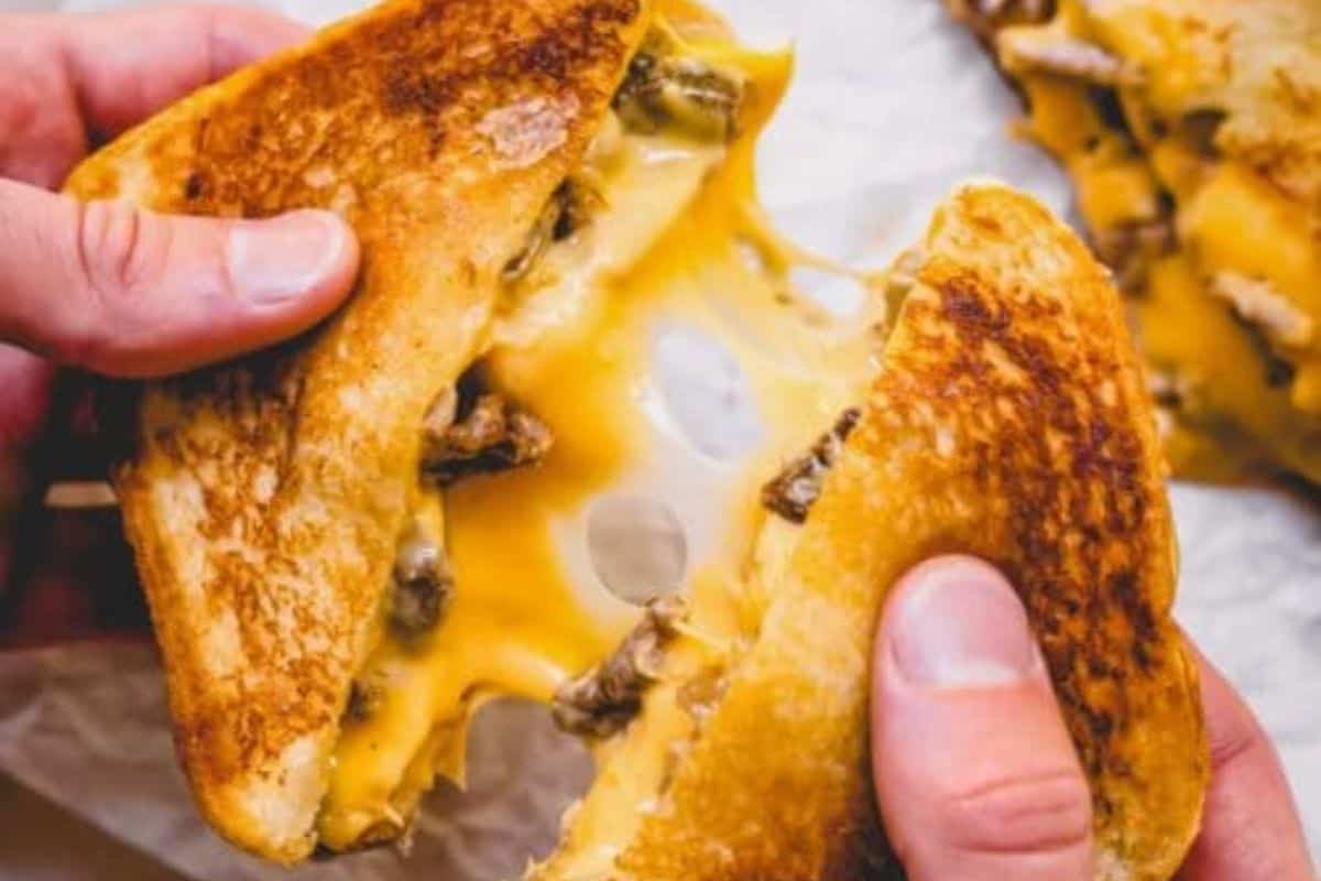  Two hands pulling apart a grilled cheese sandwich, revealing gooey, melted yellow cheese and pieces of meat or vegetables stretching between the halves.