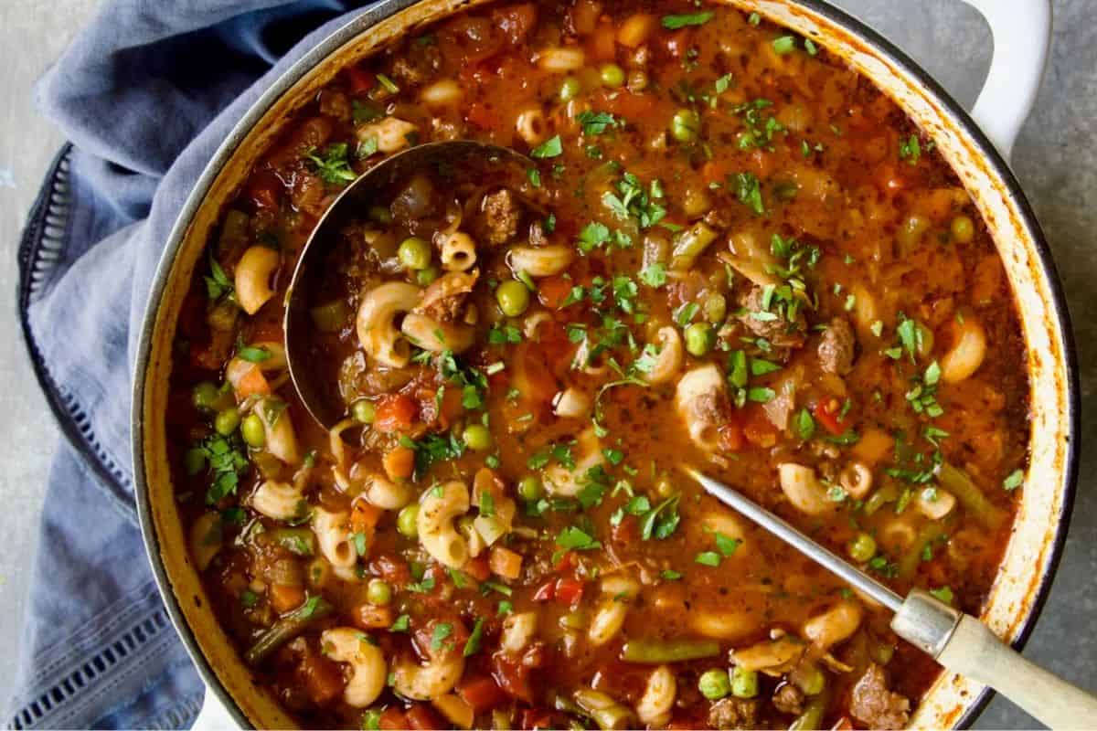 A comforting pot of vegetable and macaroni soup with ground meat, peas, beans, carrots, and tomatoes, garnished with parsley. Perfect for dinner, this hearty dish is a standout among beef soup recipes. A ladle and spoon rest in the pot.