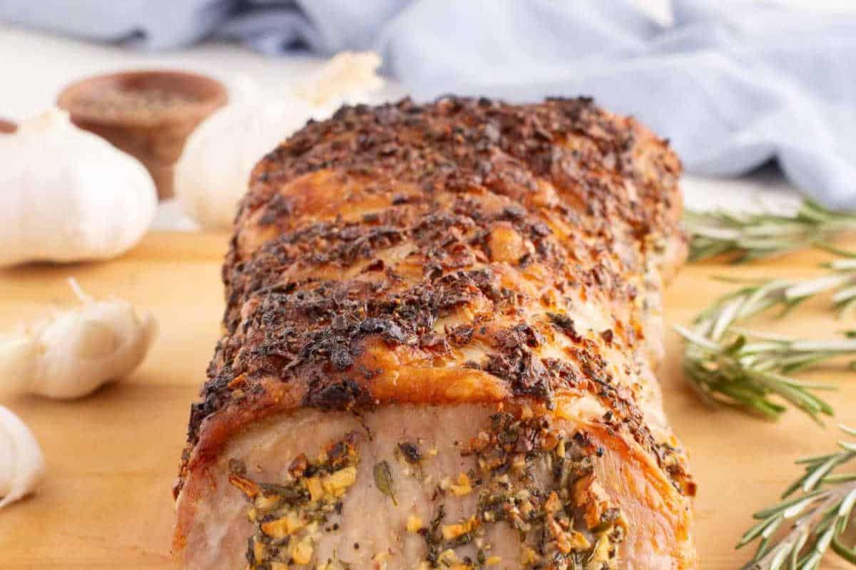 A delicious pork roast on a wooden cutting board, garnished with herbs and surrounded by garlic cloves and rosemary sprigs. Perfect for trying new pork roast recipes.