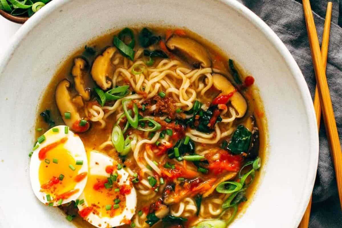 Bowl of flavorful ramen noodle soup topped with sliced mushrooms, soft-boiled egg halves, green onions, and red chili sauce, with chopsticks placed beside the bowl—perfect for those seeking easy ramen recipes.