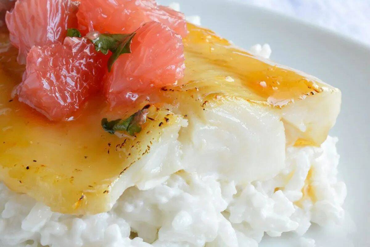 A close-up shot of a serving of honey-glazed cod fillet on a bed of white rice, topped with segments of pink grapefruit. The cod has a glossy, golden-brown glaze.