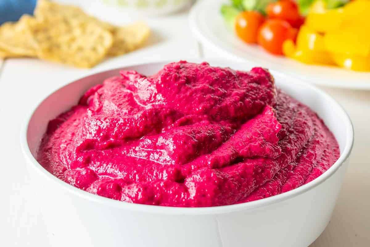 A white bowl filled with bright magenta-colored beet hummus, with a smooth, slightly textured surface. In the background, out of focus, are tortilla chips and chopped vegetables.
