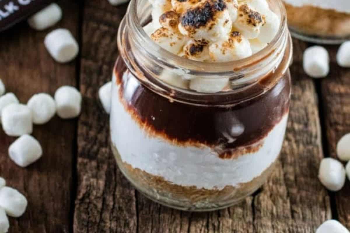  A s'mores dessert layered in a small glass jar, seen from above and slightly to the side. The layers, from bottom to top, are a light brown crumb base, a thick white marshmallow layer, a dark chocolate layer, and a top layer of small, toasted mini marshmallows. Several loose mini marshmallows are scattered on the rustic wooden surface around the jar.