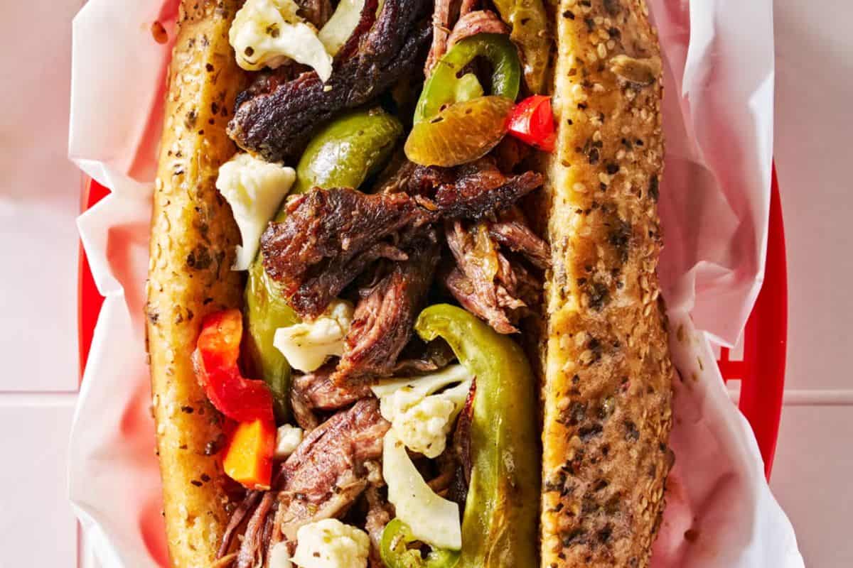 A close-up of a gourmet sandwich: an Italian beef sandwich in a seeded bun, filled with shredded beef, green peppers, pickled vegetables, and served on white parchment paper.