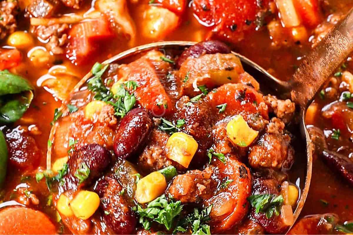 A ladle filled with comforting vegetable and bean soup featuring corn, carrots, kidney beans, and ground meat in a tomato-based broth—perfect for dinner and inspired by classic beef soup recipes, garnished with chopped herbs.