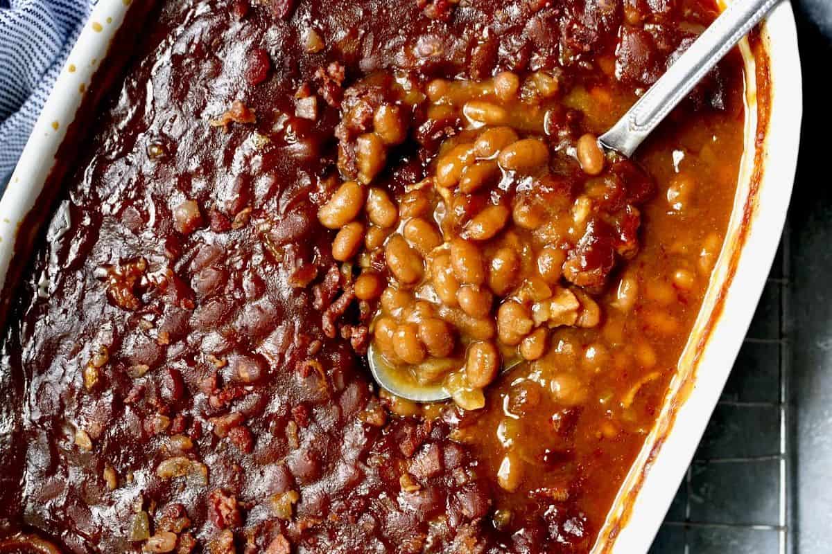A close-up of canned beans in a casserole dish with a serving spoon scooping beans from the saucy surface—perfect for delicious dishes and easy recipes.