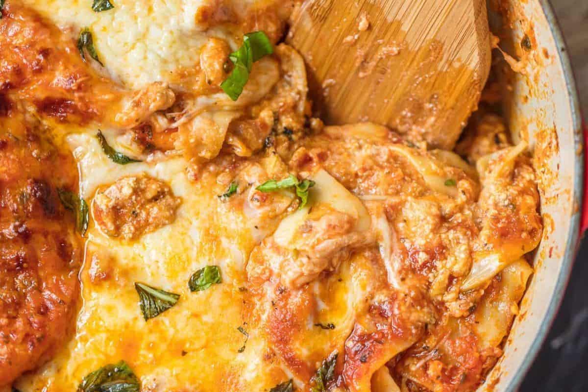 Close-up of cheesy baked lasagna with visible layers of pasta, sauce, and basil, being served with a wooden spatula—one of the ultimate hearty recipes perfect for your Dutch oven.