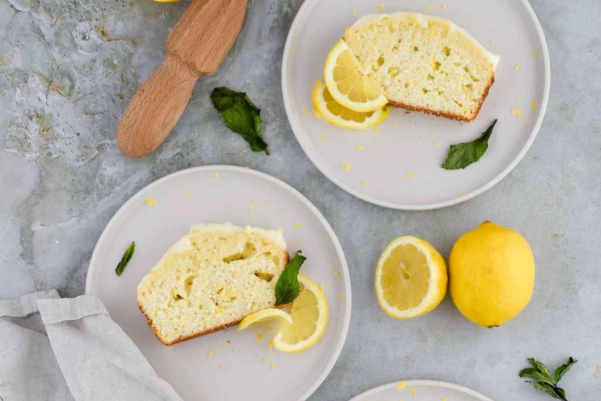 Two plates each hold a slice of lemon cake, garnished with lemon slices and mint leaves; a whole lemon and a wooden utensil are nearby, inspiring new bake recipes for quick breads on a light-colored surface.