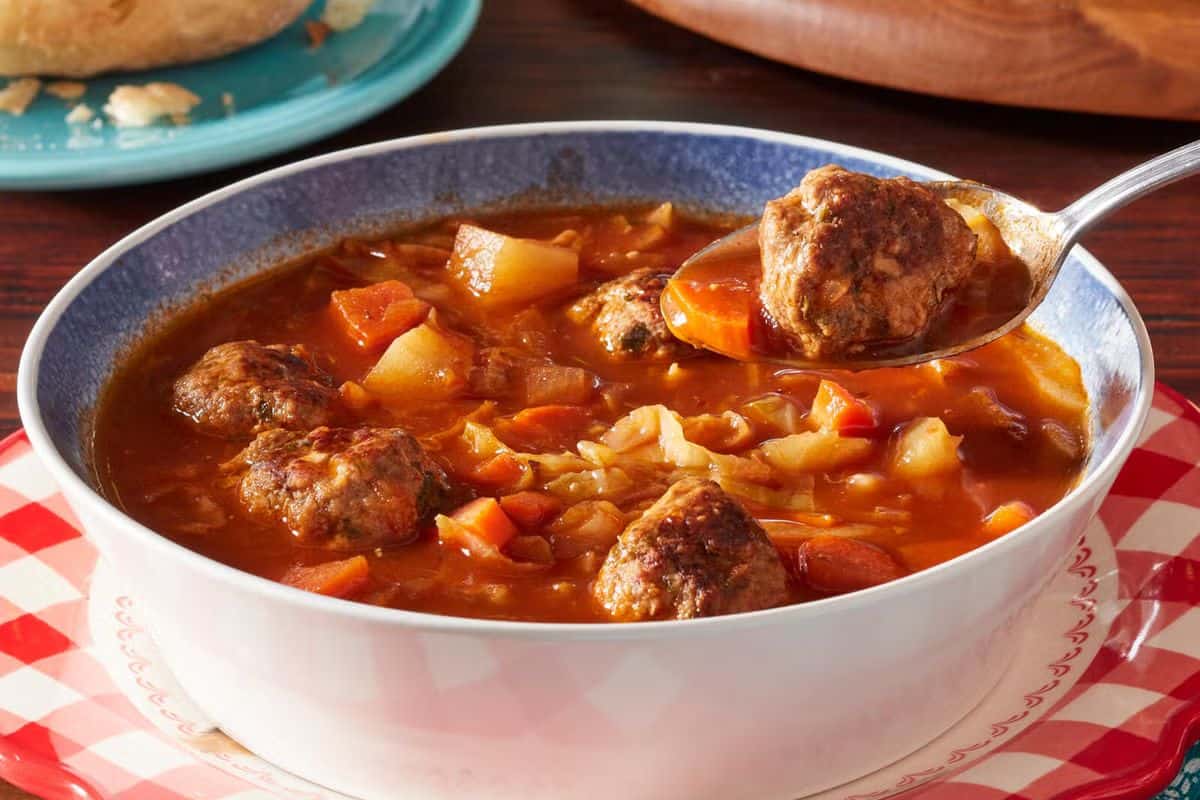 A bowl of tomato-based comforting soup with meatballs, diced carrots, potatoes, and cabbage, with a spoon lifting a meatball—perfect for your dinner rotation.