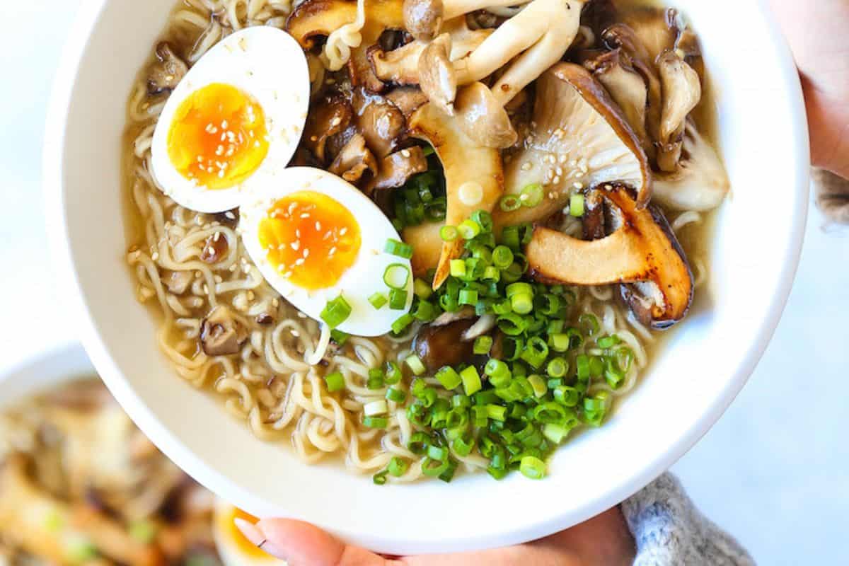 A bowl of flavorful ramen topped with sliced mushrooms, halved soft-boiled eggs, chopped green onions, and sesame seeds.