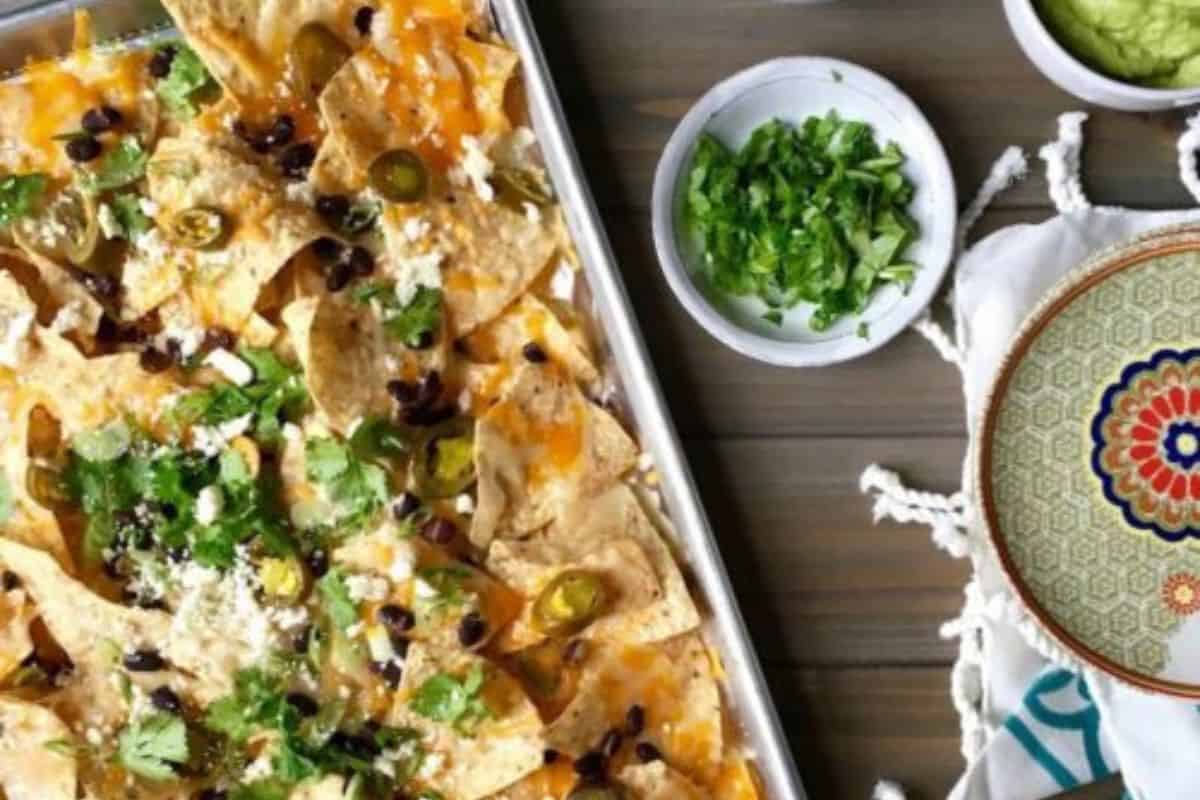 A baking sheet of nachos topped with melted cheese, black beans, jalapeños, and cilantro sits next to bowls of chopped greens and guacamole—a perfect centerpiece for party food recipes—alongside a decorative plate.