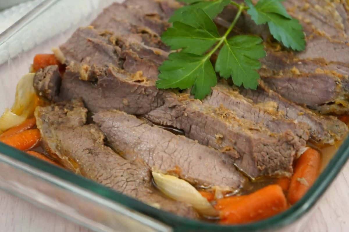 Sliced cooked brisket served in a glass dish with carrots and onions, garnished with a sprig of parsley—perfect for those who love hearty Dutch oven recipes.