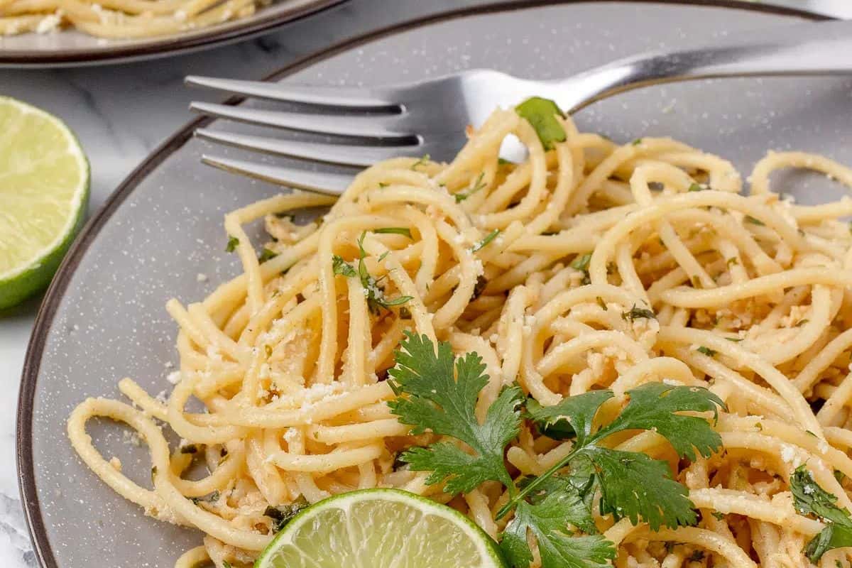A plate of spaghetti garnished with cilantro, grated cheese, and fresh lime slices, with a fork resting on the plate—perfect inspiration for your next cooking adventure or to add to your favorite recipes.