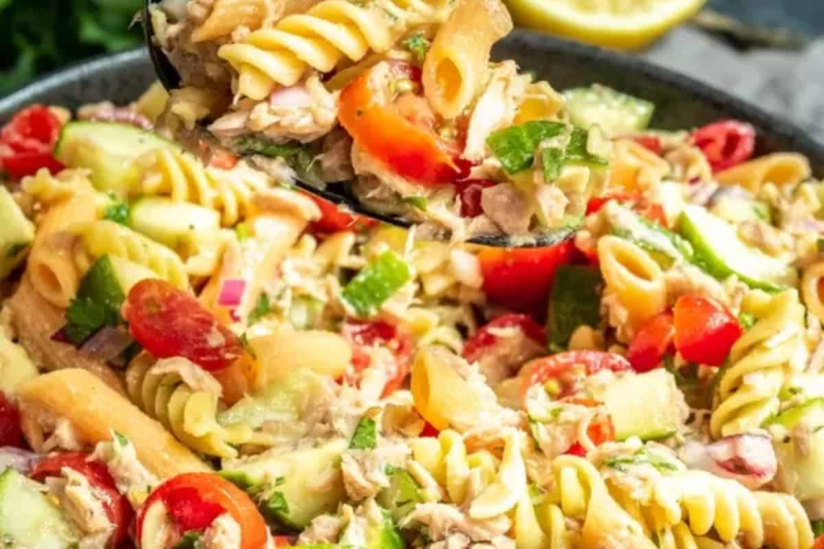 A close-up of a pasta salad with rotini, cherry tomatoes, cucumbers, red onions, parsley, and chunks of tuna in a serving spoon and bowl—perfect for easy tuna meals or anyone searching for delicious tuna recipes.