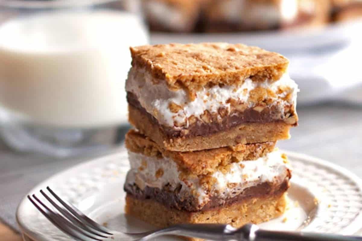 Two s'mores bars stacked on a white plate with a fork in front and a glass of milk in the blurred background. Each bar has a light brown base, a layer of dark chocolate, and a thick layer of toasted white marshmallow on top, with a light brown crumbly topping.