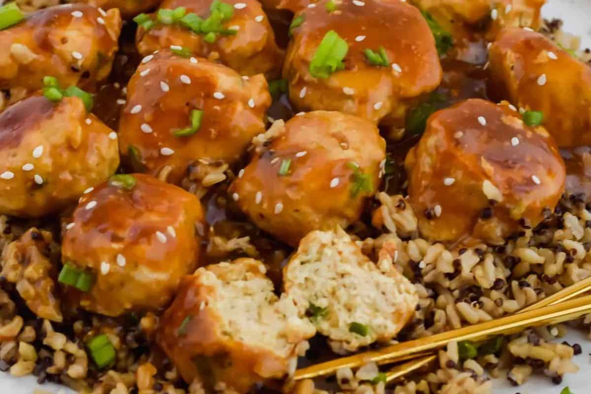 Chicken meatballs topped with sesame seeds and chopped green onions served over rice with a savory brown sauce—perfect for serving warm or as one of your cold appetizer ideas for gatherings. A fork cuts into one meatball, revealing its juicy inside.