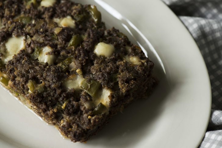 A slice of crave worthy meatloaf with visible chunks of green peppers and melted cheese, inspired by cheesesteak recipes, is served on a white plate.