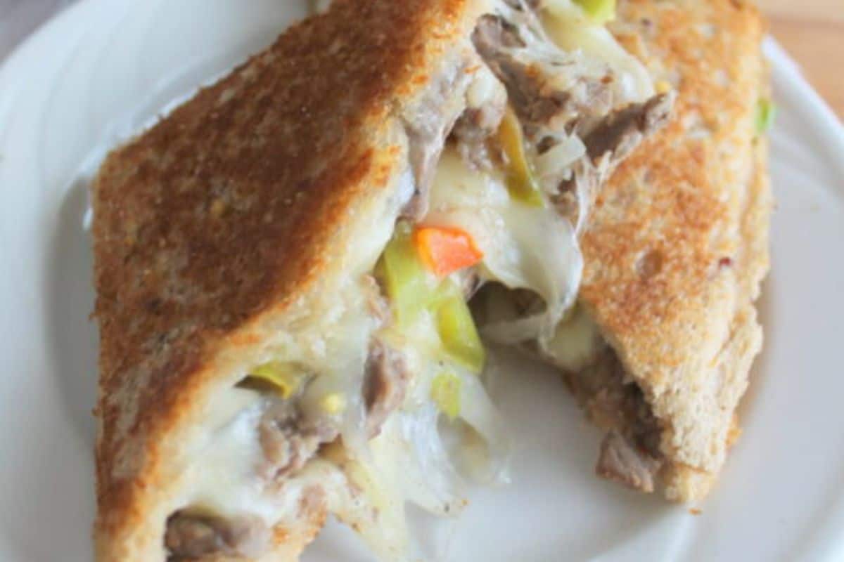 A toasted sandwich with visible melted cheese, shredded meat, and chopped green and orange vegetables (possibly bell peppers or carrots), cut in half and served on a white plate.