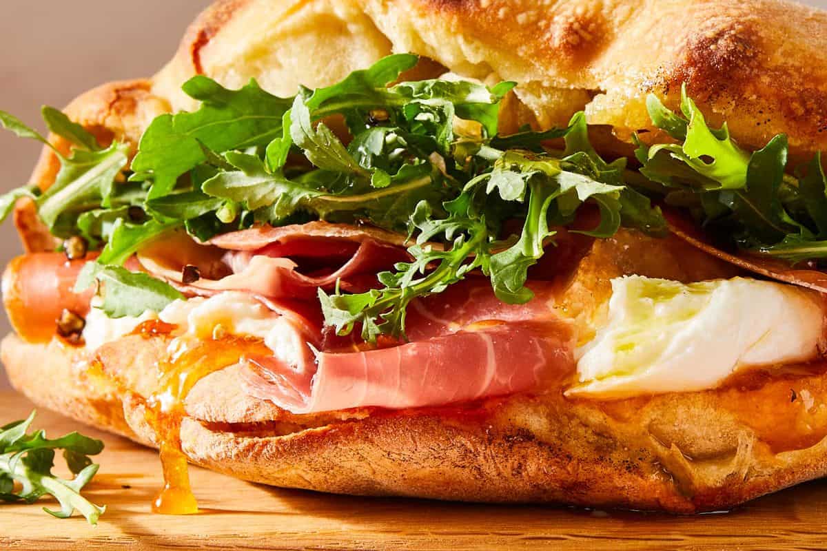 A close-up of a gourmet sandwich with prosciutto, fresh mozzarella, arugula, and a crusty bread roll on a wooden surface—perfect for lovers of gourmet sandwiches.