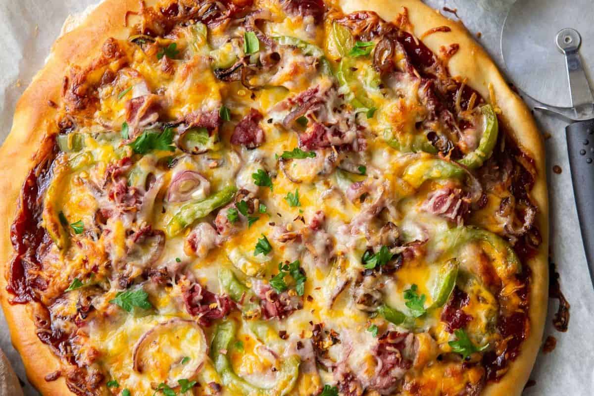 A close-up, overhead view of a whole pizza topped with melted cheese, green bell peppers, red onions, meat (possibly bacon or ham), and garnished with fresh parsley.