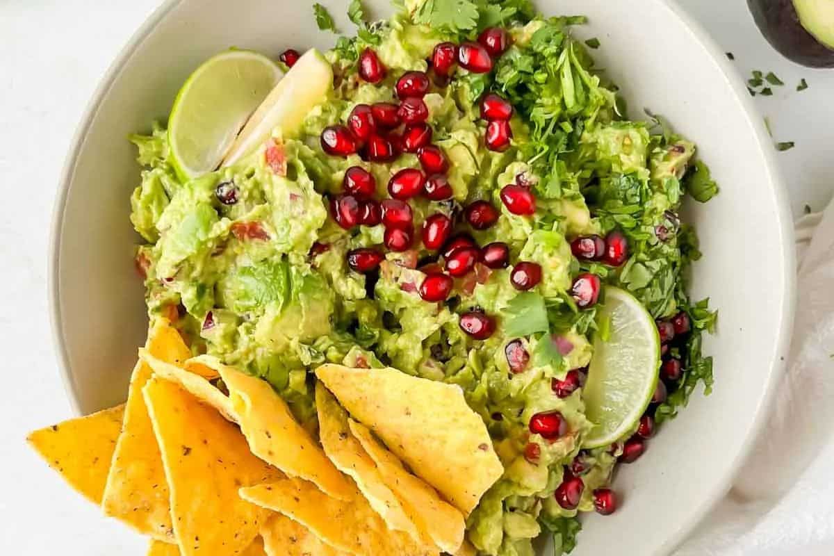 A bowl of guacamole topped with pomegranate seeds, cilantro, and fresh lime slices, served with tortilla chips on the side—perfect inspiration for new recipes and creative cooking.