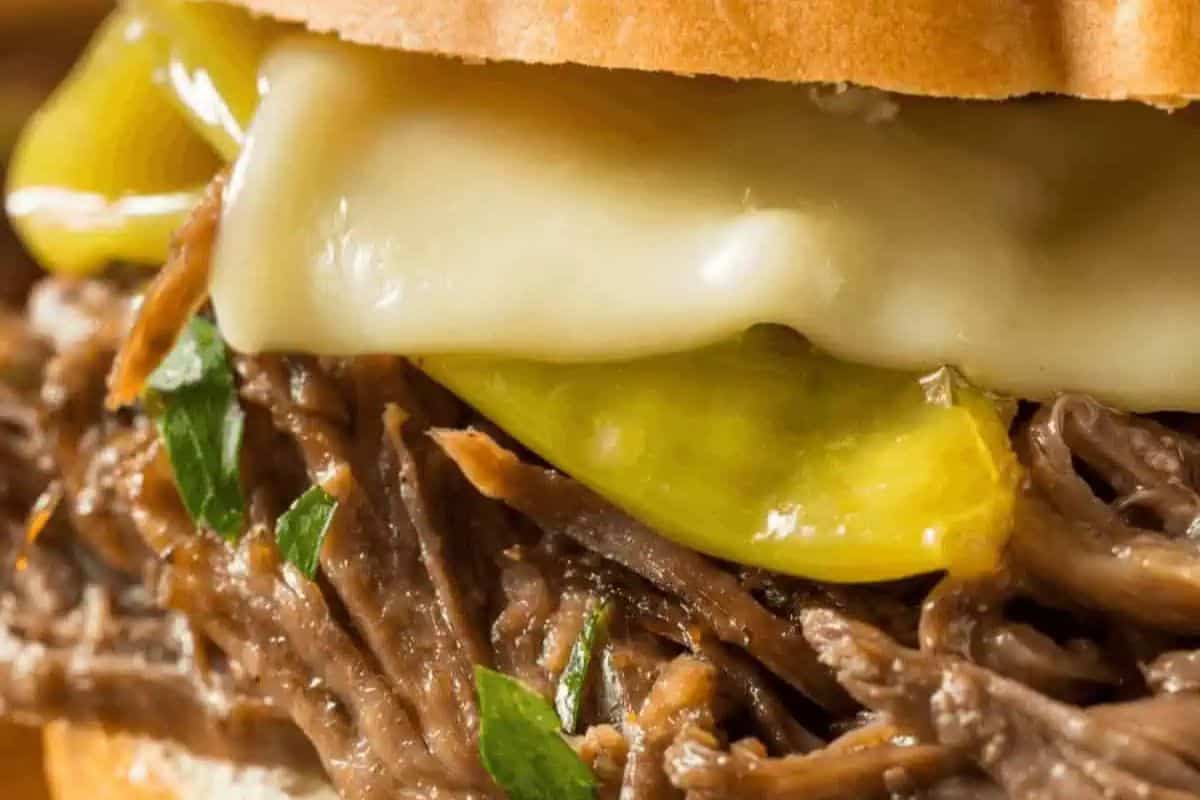 Close-up of a gourmet sandwich with shredded beef, melted cheese, sliced pepperoncini peppers, and parsley on a bun.