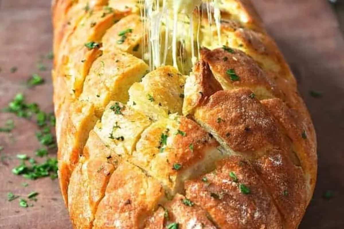 A loaf of pull-apart garlic bread with melted cheese stretches between slices, garnished with chopped parsley on a wooden surface—perfect for party food recipes.